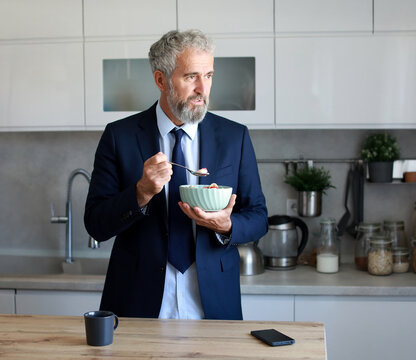 Portrait of a mid aged mature man gettyng ready for work eating breakfast oatmeal in a bowl standing in kitchen home. Overworked, busy, stress, urgency, multitasking, active healthy senior  concepts