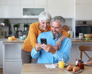Portrait of an elderly senior couple having breakfast and using a smart phone at home. Happy healthy affectionate senior couple talking and showing smartphone screen to one another and texting and sit