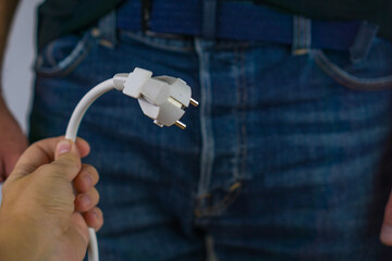 hand holding a white european power plug with a man in blue jeans and a dark shirt in the background concept of male sexual health, energy and power, erectile dysfunction