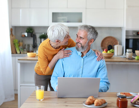 Portrait of an elderly senior couple having breakfast and looking at a laptop at home. Happy healthy affectionate senior couple eating and sitting at kitchen table having fun enjoying morning meal tog