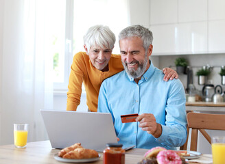 Portrait of an elderly senior couple having breakfast and looking at a laptop using a credit card for online shopping at home. Happy healthy affectionate senior couple eating and sitting at kitchen ta