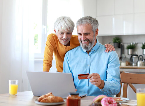 Portrait of an elderly senior couple having breakfast and looking at a laptop using a credit card for online shopping at home. Happy healthy affectionate senior couple eating and sitting at kitchen ta - Powered by Adobe