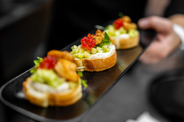 Gourmet bruschetta with creamy spread, microgreens, diced vegetables, seafood, and red roe on toasted bread, served on a black tray, held by hand. Elegant appetizer presentation.