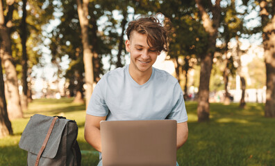 Fototapeta premium Cheerful student guy with laptop computer doing homework or attending online lecture, sitting on bench outdoors. Distance education concept