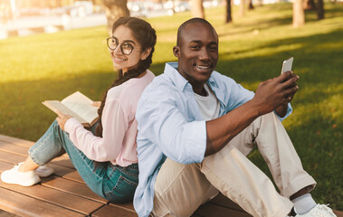 Happy diverse college students sitting back to back, using smartphone and book, resting outdoors on...