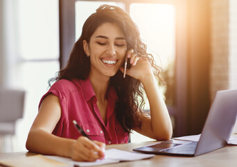 A woman with long, dark hair sits at a desk, smiling as she talks on the phone. She is taking notes while working on a laptop in a sunlit room.