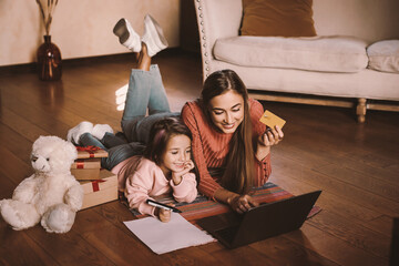 Online Shopping For Xmas Eve. Cheerful female using pc and debit card, preparing for holiday together with little daughter lying on floor writing check list of festive presents in living room with pen