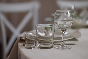 Three empty glasses of different shapes on a beige tablecloth in a softly lit dining setting with white chairs. Concept hospitality, event catering, table setting, restaurant service.