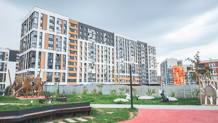 Modern residential courtyard with wooden playground, benches, green lawn and decorative sculptures surrounded by high-rise apartment buildings in orange, white and gray tones under cloudy sky