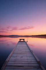 Obraz premium A serene sunrise paints the sky in pink hues over a tranquil lake, reflected perfectly on the waters surface. A wooden dock extends into the calm water, promising peace and quiet.