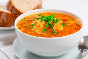 A bowl filled with bright orange soup garnished with green chives sits on a white table. Fresh slices of brown bread accompany the bowl, adding a comforting touch to the meal