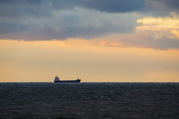 A solitary cargo ship navigates a stormy sea under a dramatic cloudy sky with a warm sunset glow on the horizon.