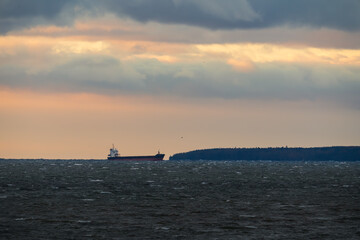 Obraz premium An industrial freight ship navigates the stormy Baltic Sea by Naissaar Island, Estonia, against a beautiful, cloudy sunset.