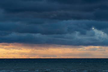 Powerful storm clouds loom over the rough Baltic Sea, with a dramatic contrast of golden sunset light on the horizon.