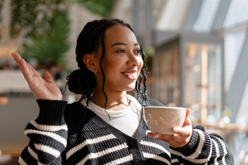 Smiling authentic woman with braids and piercing enjoying coffee in cafe waving hand