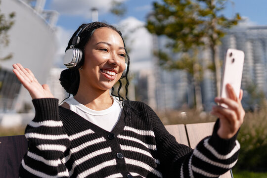 Young smiling African American woman having video call on smartphone in park waving hand - Powered by Adobe