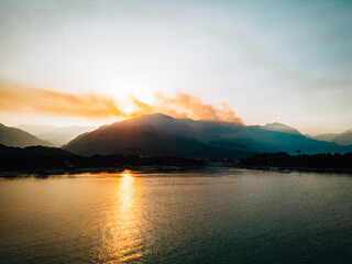 Drone view of sunset with smoke over mountain peaks and golden light reflecting on calm sea. Dramatic landscape with smoky sky, boats, and coastal silhouettes