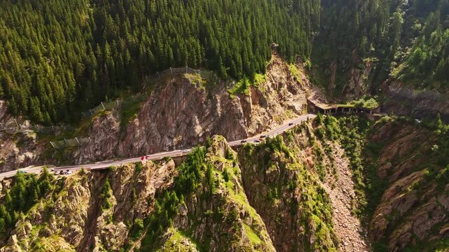 Cars ride by the highway in the spectacular high rocks. Pine tree woods cover the mountain slopes. Wild nature of Romania, USA. Drone footage.