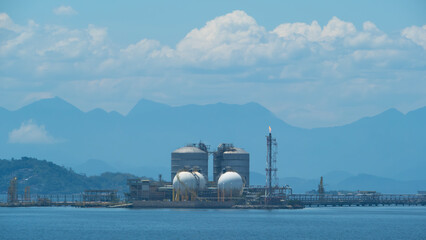 Industrial complex with large storage tanks on a pier, set against a backdrop of mountains and a...
