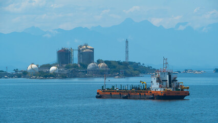Bunker barge at anchor in calm blue waters near an industrial facility with storage tanks, mountains visible in the distance © John_Doo78