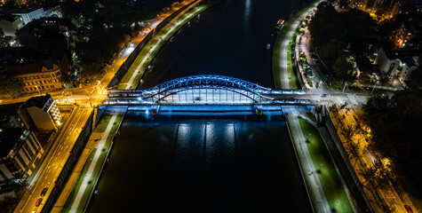 Tytuł: Night aerial view of a blue steel bridge over the Vistula River in Krakow Opis: Stunning nighttime drone view of a blue illuminated steel bridge over the Vistula River in Krakow, Poland. Bright © Sawomir