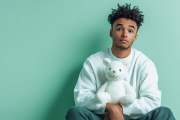 Thoughtful young man embracing a white teddy bear, feeling emotional support and well being on a mint green background