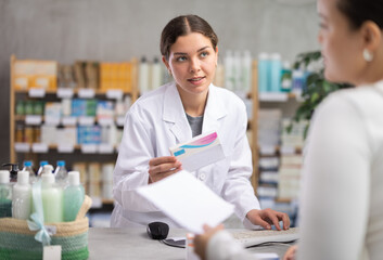 Obraz premium Smiling friendly young female pharmacist in white coat assisting woman, visiting pharmacy, selecting medications and checking availability through computer system