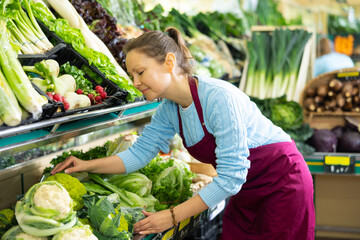 Smiling middle-aged European woman in apron selling cauliflower inflorescence in grocery shop