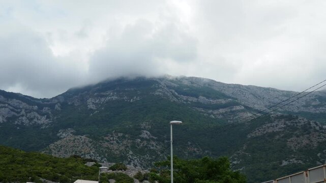 Clouds embrace the peaks of the mountains while lush greenery flourishes at the base. A rural landscape is visible, with structures dotting the scenery. Montenegro, Canj, Bar