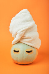 A pumpkin with eyes and a white terry towel on its head, a blue clay mask on its face, an orange background