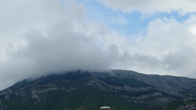 Clouds roll over the rugged mountain, creating a captivating view. The lush greenery contrasts with the grey rock faces under the vast sky. A tranquil moment in nature unfolds. Montenegro, Canj, Bar
