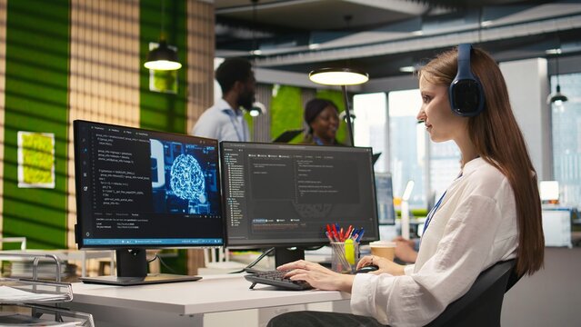 Young female programmer in office reviewing AI system metrics, listening music, monitoring performance dashboards. Junior dev wearing headphones tuning neural networks using tools on PC, camera A - Powered by Adobe