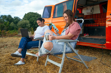 Women relaxing on chairs working with laptop and phone outside camper van.