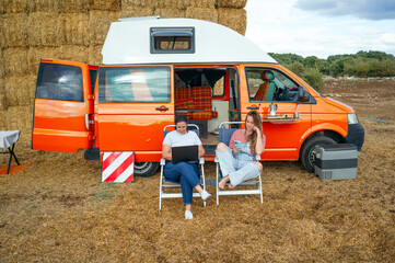 Women relaxing on chairs working with laptop and phone outside camper van.