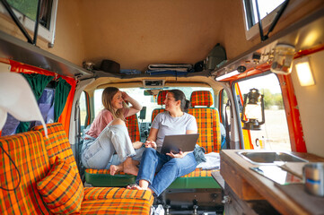 : Two women relaxing inside a camper van, sitting on orange plaid seats, talking and working on a laptop.