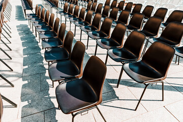 Rows of black chairs are neatly arranged on a paved surface, casting crisp shadows in the daylight. Empty outdoor chairs set up in neat, receding rows, casting distinct shadows on the stone flooring