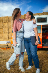 Two women laughing and hugging outdoors beside an orange camper van and hay bales.
