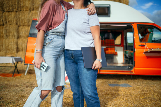 Two women laughing and hugging outdoors beside an orange camper van and hay bales.