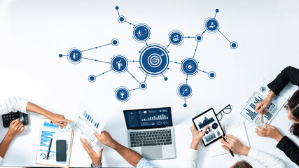 Overhead view of a collaborative meeting where team members analyze data on laptops and tablets, connecting insights through blue graphic icons to achieve business goals. Amity