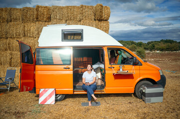 Woman relaxing barefoot with coffee outside orange camper van in rural area.
