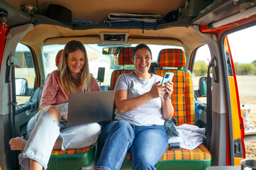 Two women sitting inside camper van smiling using laptop and smartphone