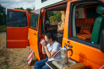 Woman sitting on camper van step drinking coffee from metal cup outdoors.