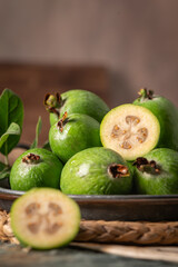A close-up, moody still life of a metal bowl filled with green feijoa, with one fruit sliced in half, on a rustic wooden table