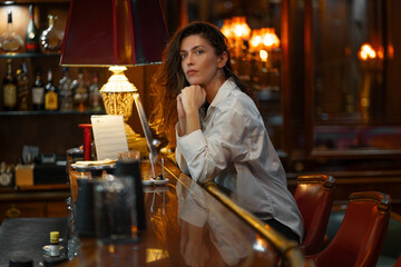 Young woman in white shirt resting her chin on hand at bar counter. Soft lighting creates warm, intimate atmosphere with bottles and vintage lamp in luxury hotel