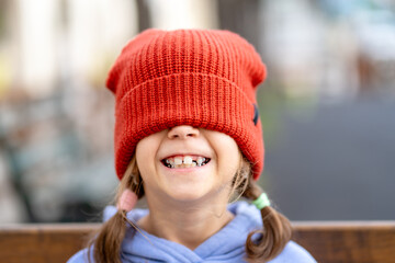Cheerful girl with red knit cap covering eyes smiling outdoors. Child with sunlight on face against blurred natural background. Closeup portrait