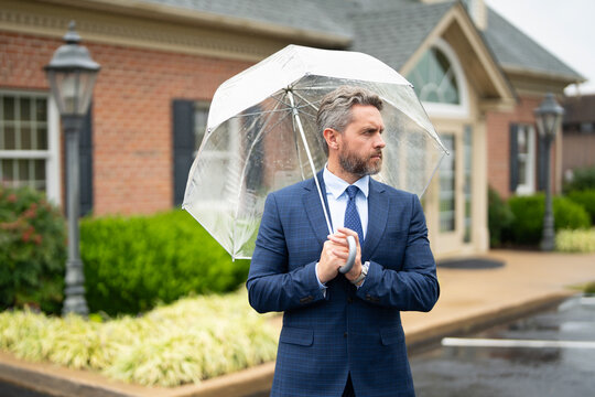 A well-dressed businessman waits patiently on a rainy street. An executive stands outdoors with an umbrella during a rain day. A successful businessman waiting near the office.