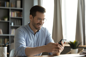 Joyful young male office employee sitting at desk reading phone