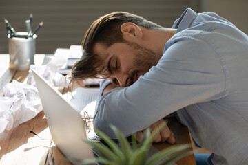 Fatigued exhausted young man freelancer sleeping on messy work desk