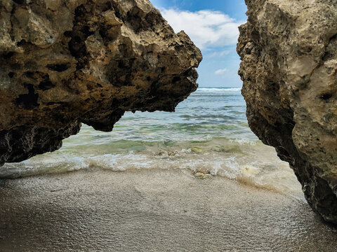 View of ocean waves framed between rugged coastal rock formations on a sandy beach under a bright, cloudy sky