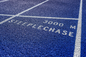 Close up photo of outdoor blue running track with white lane lines, with the word, steeplechase 3000 M, on a new track.